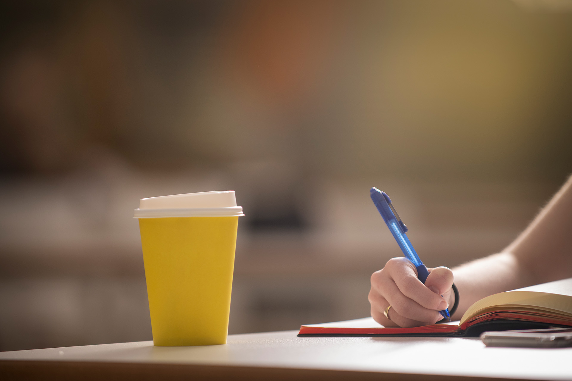 Disposable yellow coffee cup on a desk while a student is taking notes in a notebook.