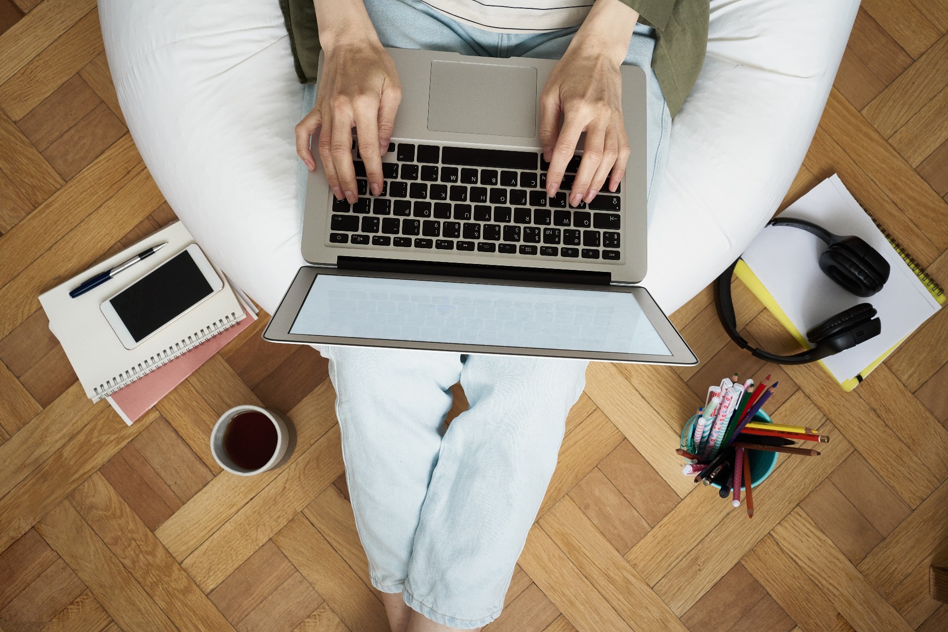 Shot from above, a person is sitting on a bean bag and typing on a laptop. The bean bag is surrounded by stationery, notebooks and a cup of cofee. 