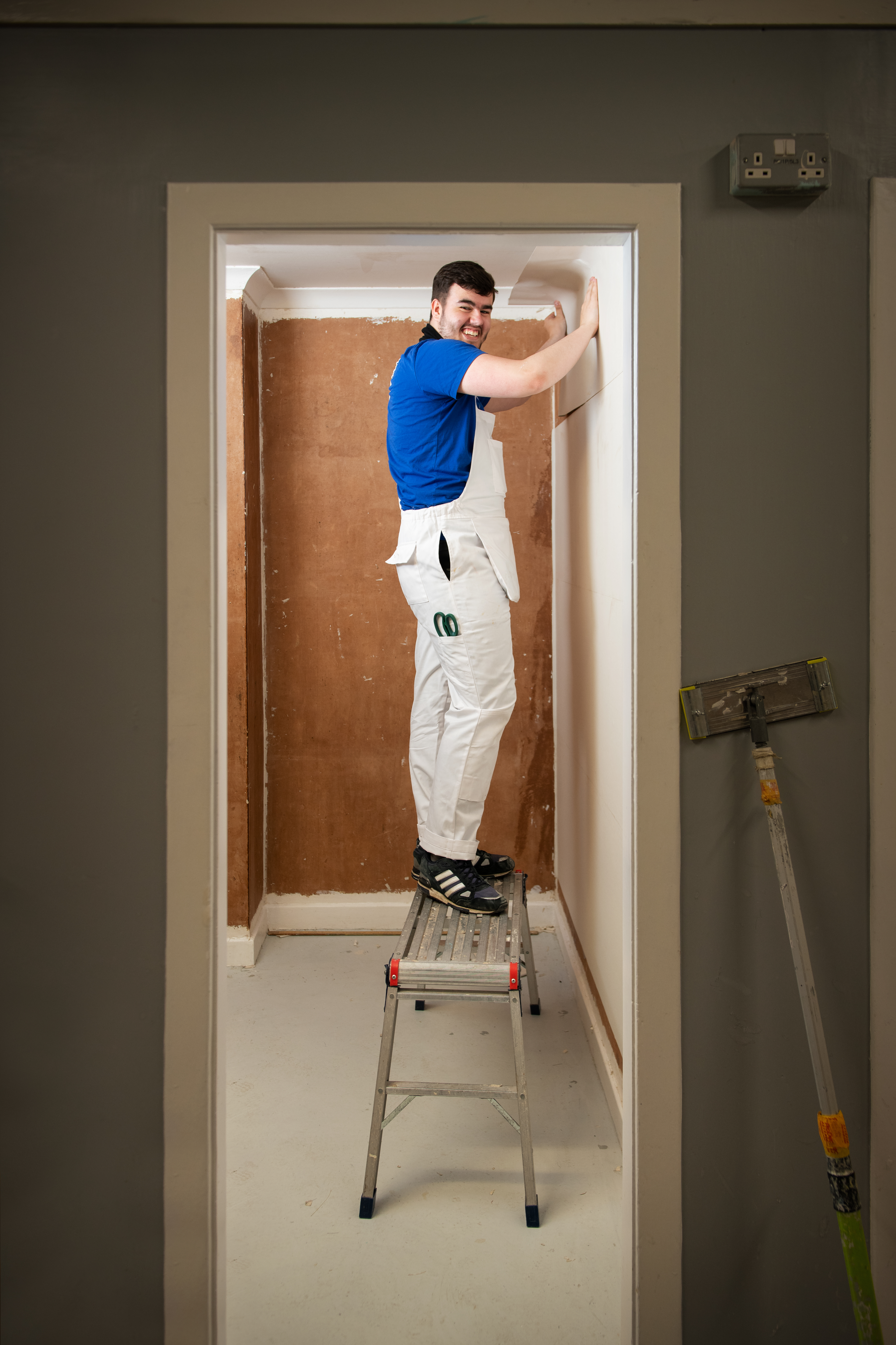 A person stands on a step ladder, applying finishing touches to a wall in a partially renovated room with a bare, brown wall.
