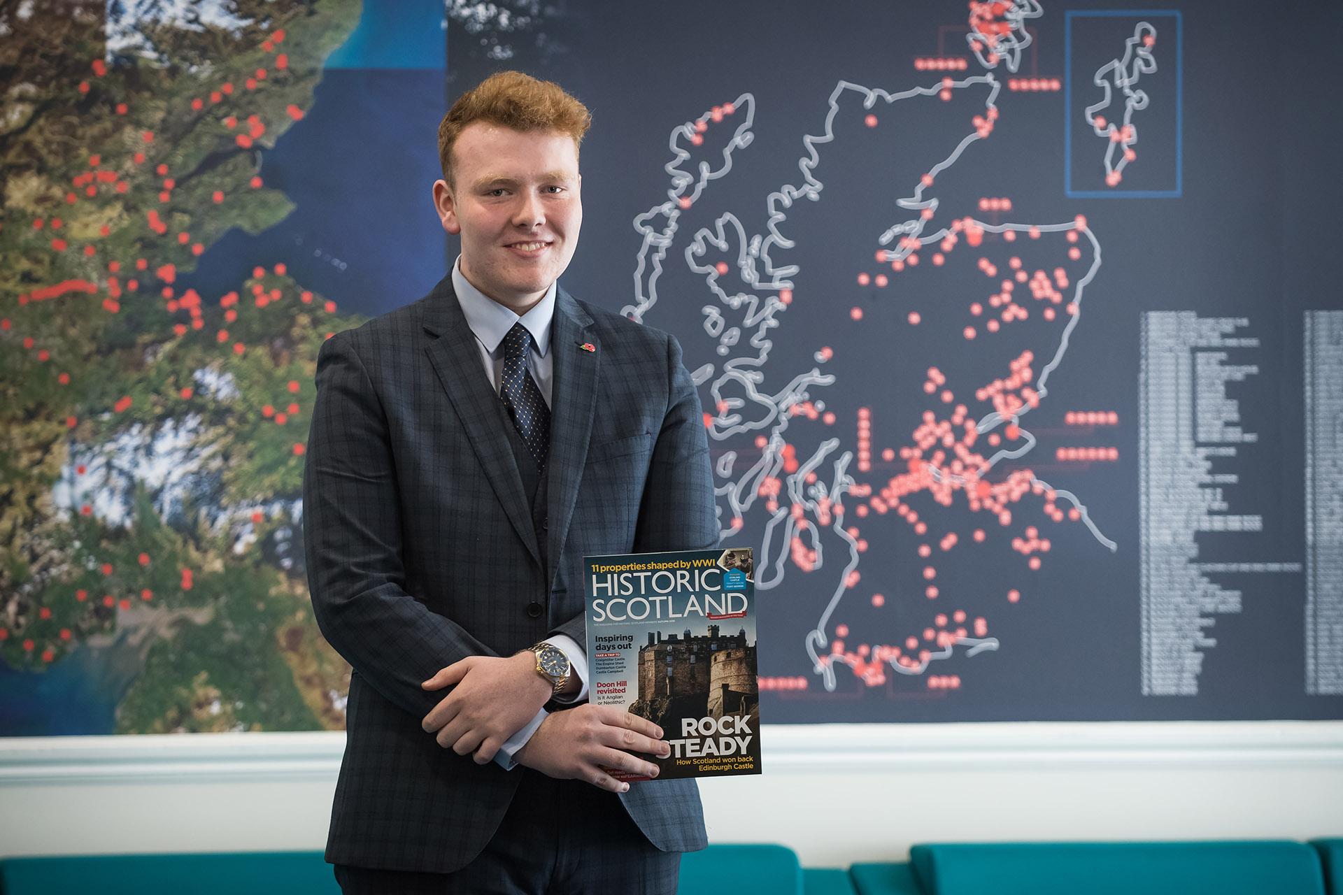Man wearing a suit, holding an Historic Scotland magazine infront of a board featuring a map of Scotland with different locations marked by red dots.