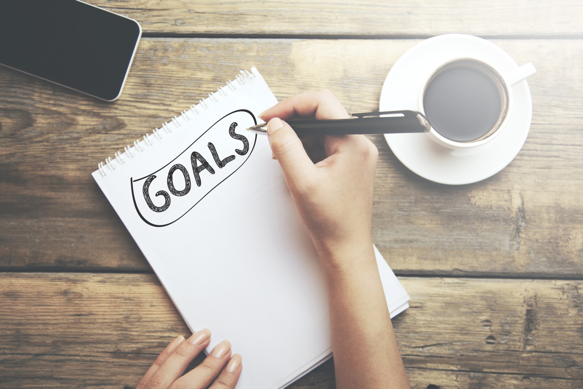 Notebook on a table surrounded by a mobile phone and cup of coffee. The word "Goals" is written on the notebook page.