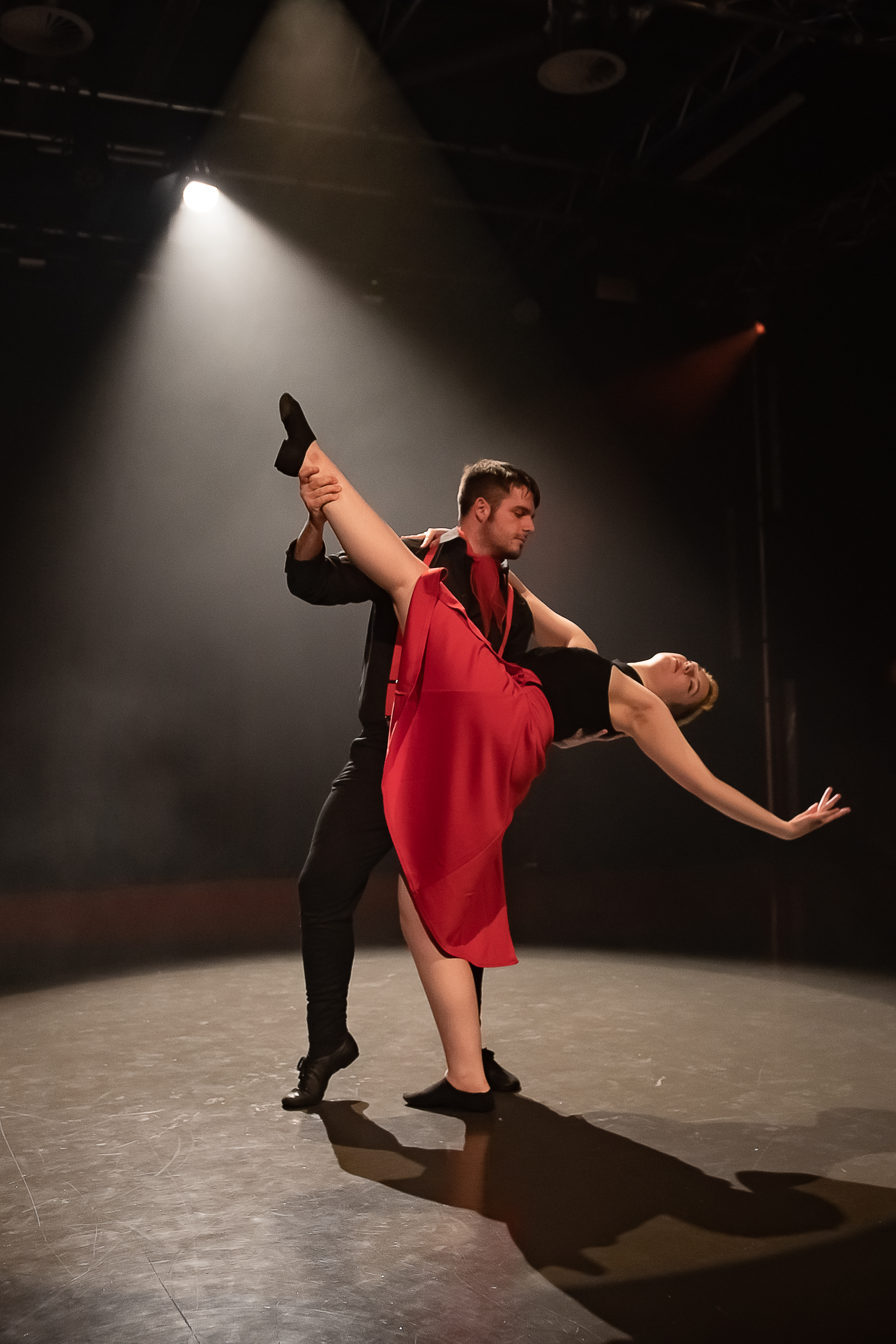 Two dancers perform on stage; the man supports the woman as she leans back with one leg lifted, both wearing black and red costumes under dramatic lighting.