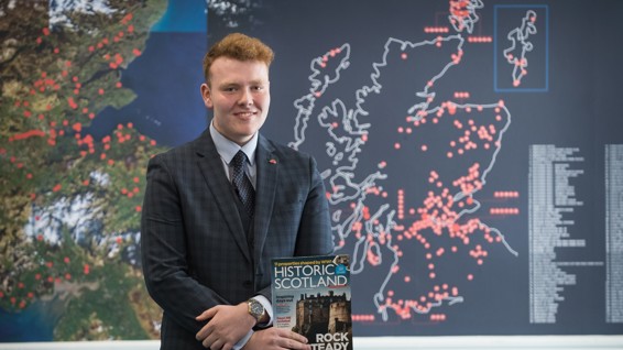 Young student wearing a suit, holding a Historic Scotland magazine at the front of a classroom. 