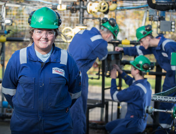 Oil and gas student smiling on site, wearing a blue boiler suit, hard hat and safety goggles.
