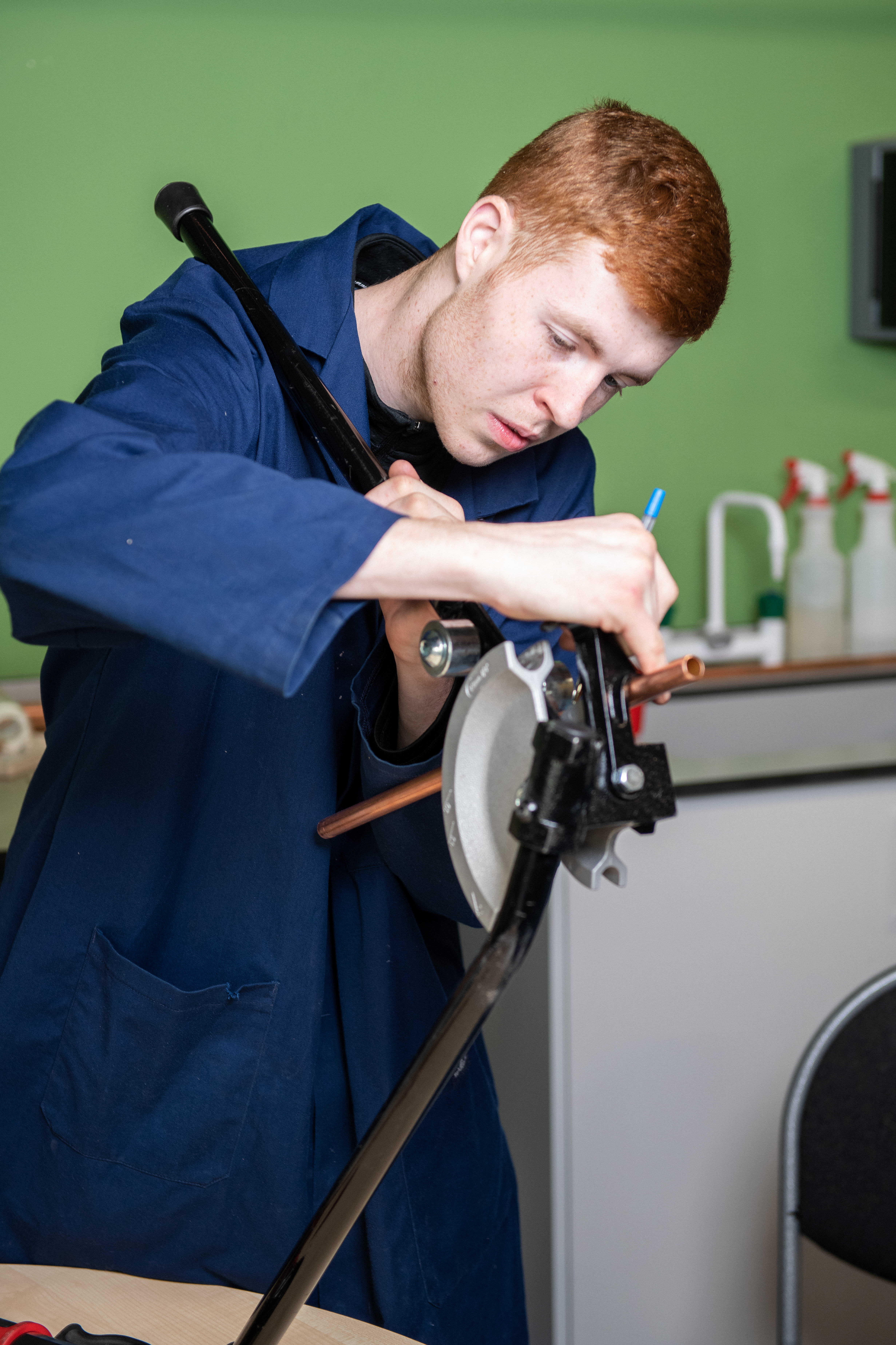 A person in a blue work jacket is bending a copper pipe with a pipe bender tool, focusing intently on the task.