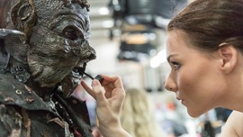 A character in an elaborate mask and textured makeup, resembling a creature sits while someone applies finishing touches with a brush.