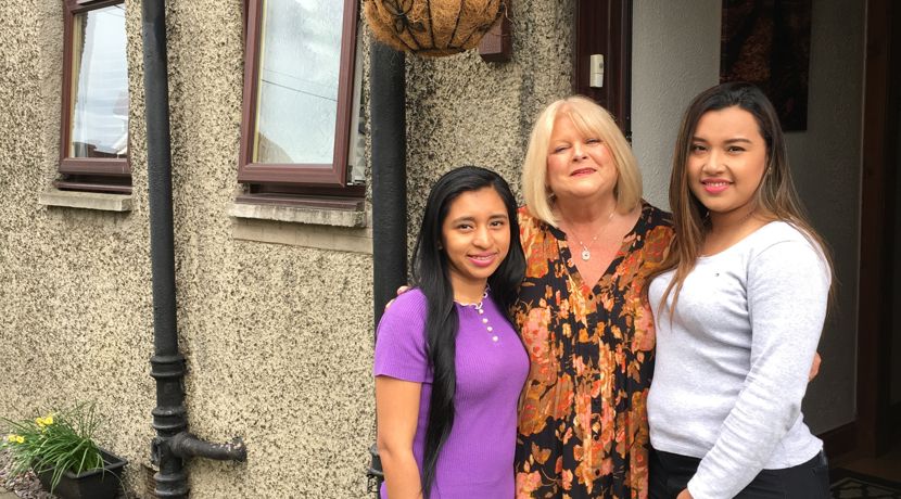 Three women stand together outside a textured house, with a hanging plant above and garden accessories visible on the ground.